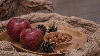 Elma ve fındıkla dolu bir sonbahar. Sonbahar arkaplanı elmalarla örülmüş sıcak bir eşarp, ahşap bir tabak, sonbahar yaprakları, fındık ve koni.