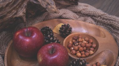 Elma ve fındıkla dolu bir sonbahar. Sonbahar arkaplanı elmalarla örülmüş sıcak bir eşarp, ahşap bir tabak, sonbahar yaprakları, fındık ve koni.