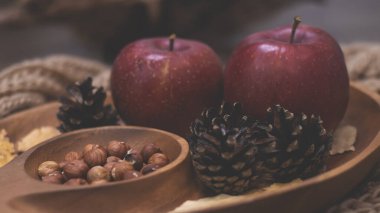 Elma ve fındıkla dolu bir sonbahar. Sonbahar arkaplanı elmalarla örülmüş sıcak bir eşarp, ahşap bir tabak, sonbahar yaprakları, fındık ve koni.