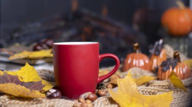 Yumuşak, örgü bir eşarp, sonbahar yaprakları, balkabağı arka planında çay olan kırmızı kupa. Sonbahar arka planı, rahat bir akşam, şömine..