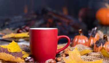 Yumuşak, örgü bir eşarp, sonbahar yaprakları, balkabağı arka planında çay olan kırmızı kupa. Sonbahar arka planı, rahat bir akşam, şömine..