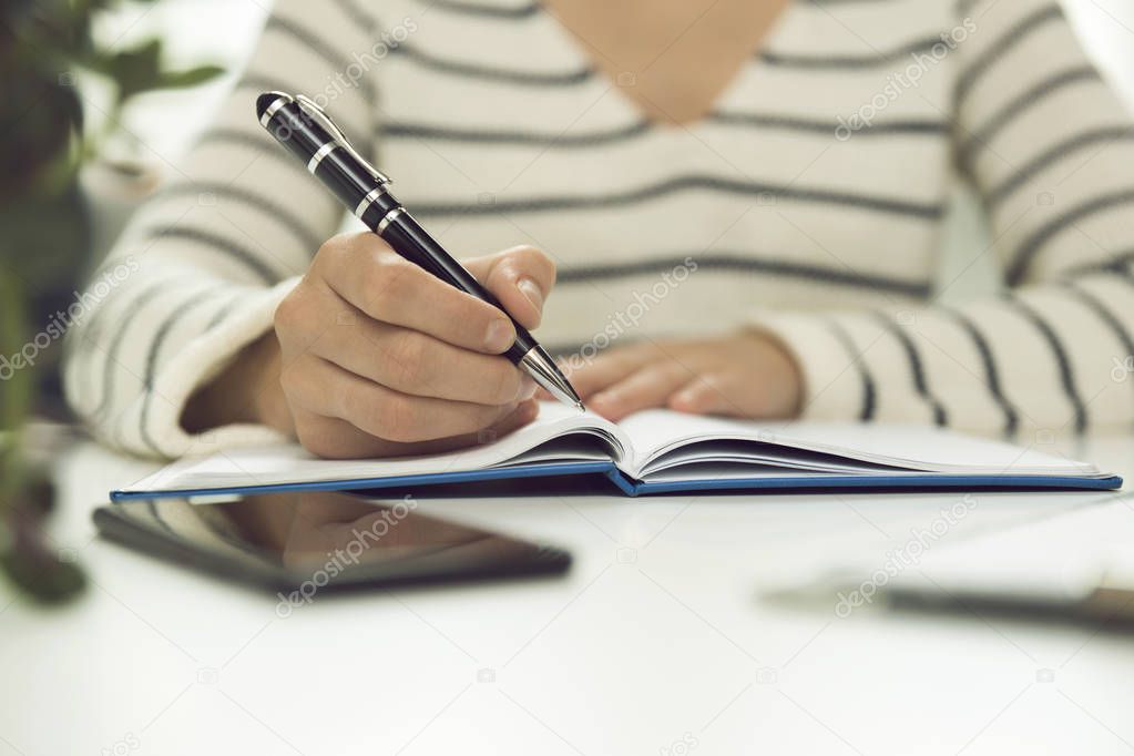 Young business woman sitting at table and writing in notebook. On table is smartphone, and tablet. Freelancer working, writing down new ideas