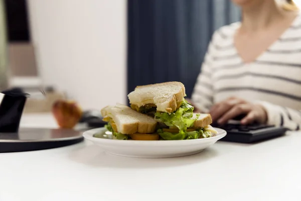 Woman eating sandwich while working. Sandwich from fresh toast with cheese and salad on work place. Work lunch