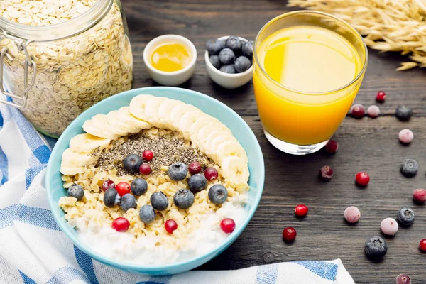Healthy fitness breakfast with fresh orange juice: oatmeal with bananas, blueberries and cranberries, chia seeds and yogurt on dark wooden board with texture