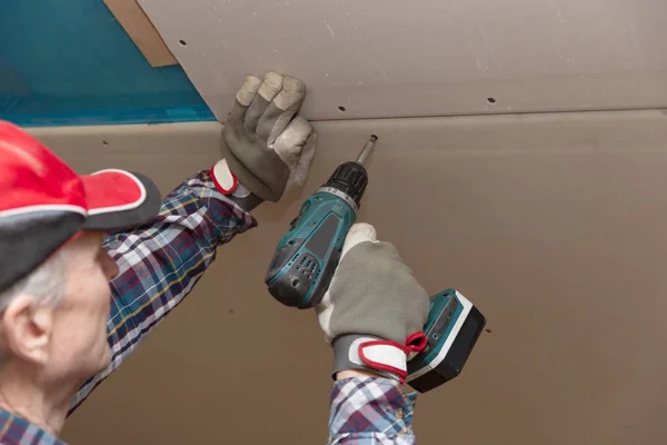 Drywall construction, attic renovation. Man fixing drywall suspended ceiling to metal frame using electrical screwdriver