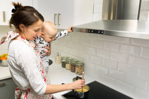 Elleri üzerinde bir bebekle genç anne aşçılar bir tencerede gıda üzerinde