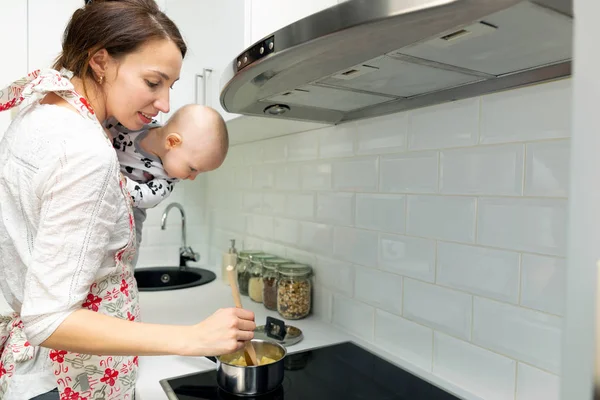 Молода мати з дитиною на руках готує їжу в горщику на — стокове фото