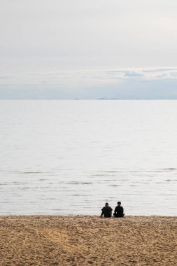 Genç erkek çift bulutlu bir öğleden sonra sahilde oturuyor ve ufukta şehir ve tekne var..