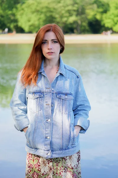 Authentic young woman wearing denim jacket over summer dress standing by lake — Stock Photo, Image