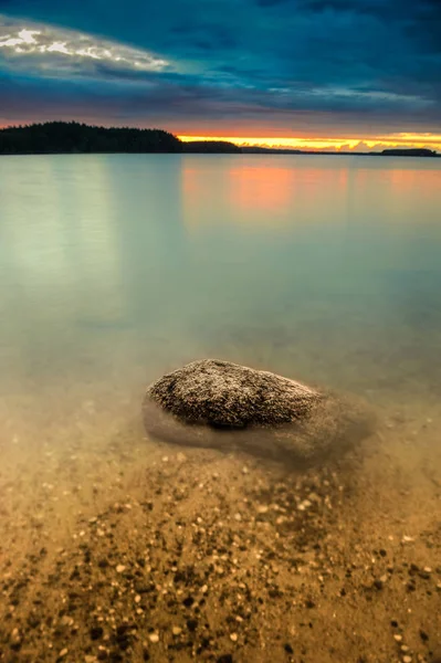 Vertical view on the lake with dark clouds on the sky and big smooth stone in the front during sunset.