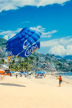 Puerto Vallarta / Mexico - Jun 26.2006: Summer view on the sandy beach and a man standing on the beach attached to the big blue parachute. People resting on the beach, sunny day.  clipart