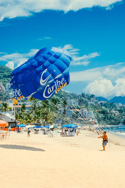 Puerto Vallarta / Mexico - Jun 26.2006: Summer view on the sandy beach and a man standing on the beach attached to the big blue parachute. People resting on the beach, sunny day. 