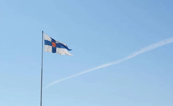 State flag of Finland flies from a flagpole against a light blue sky with a single contrail
