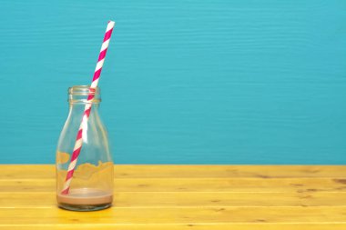 Üçte bir bardaklık süt şişesi eski bir kağıt kamışla karışık çikolatalı milkshake ile ahşap bir masanın üzerinde, deniz mavisi zeminine karşı.