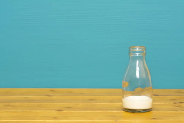 One-third pint glass milk bottle with dregs of fresh creamy milk, on a wooden table against a bright teal painted background