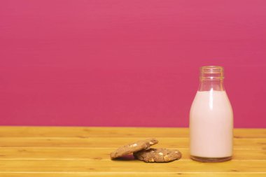 Çilekli milkshake, yarım litre bardakta süt şişesi ve çikolata parçalı kurabiye, ahşap bir masada, pembe boyalı arka planda.