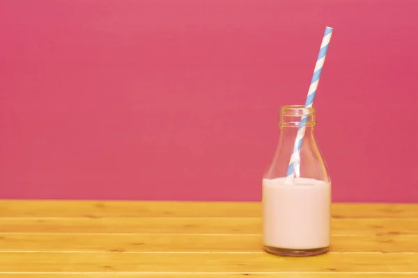 One-third pint glass milk bottle half full with strawberry milkshake with a retro paper straw, on a wooden table against a pink background