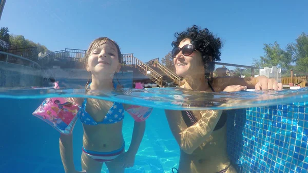 Family, mother and daughter relaxing sitting on stairs in luxury pool with spa thermal water,  half underwater shot