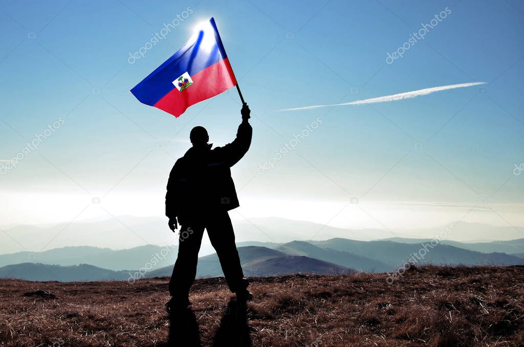 silhouette of man holding waving Haiti flag on top of mountain peak