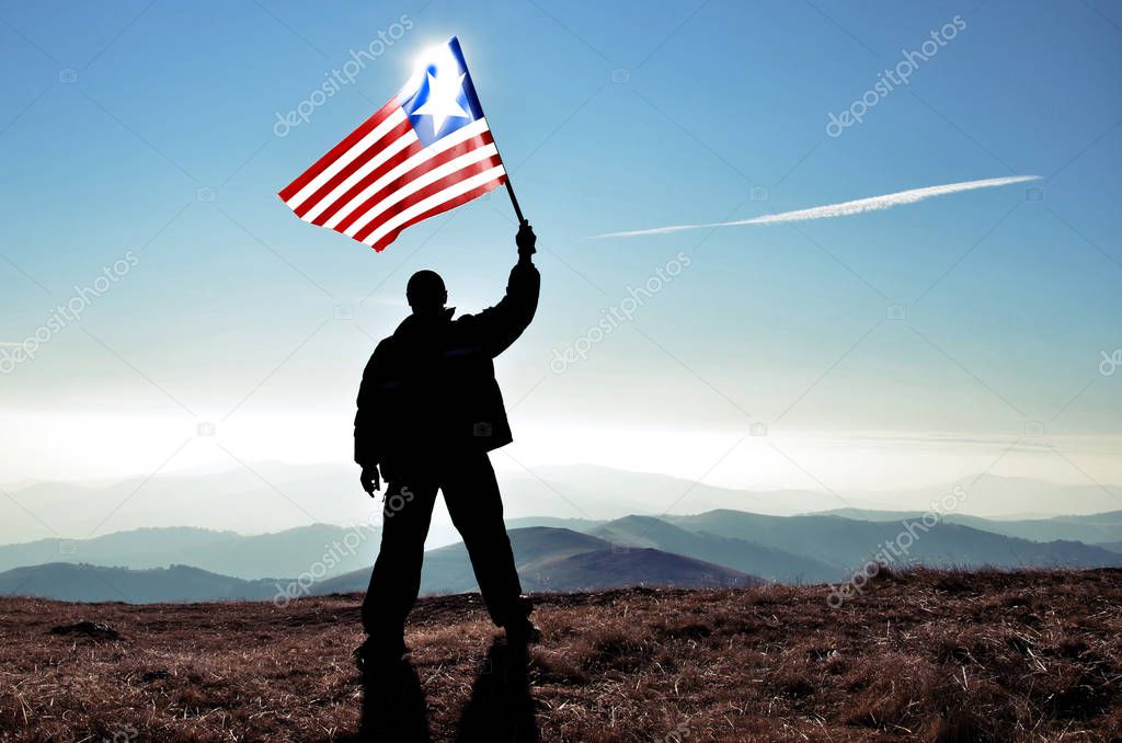 silhouette of man holding waving Liberia flag on top of mountain peak
