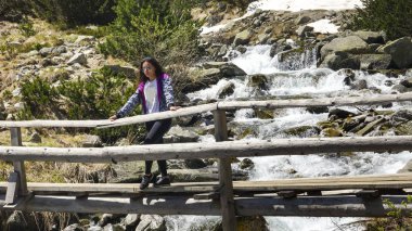 Sport girl hiking and enjoing on old wooden bridge the fast flowing waters from snow melting during springtime high on mountains clipart