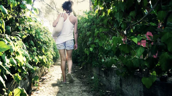 Rear view of woman in shirt and slippers walking to beach on narrow foliage passage