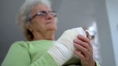 Yaralı elini bandajla sararak oturan üzgün yaşlı kadın.