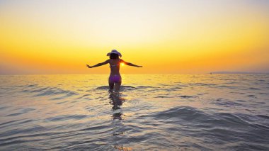 Bikinili ve şapkalı seksi genç gün batımında deniz suyundan çıkıyor, yaz tatili konsepti.