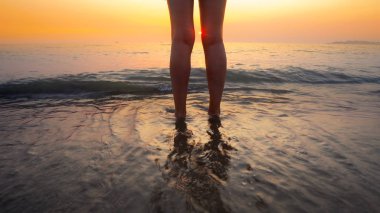 Sahilde gün batımında kadın ayaklarına su sıçrattı. Huzurlu ve rahatlatıcı okyanus suyu sahnesi güzel sahilde ayaklara sıçrıyor.