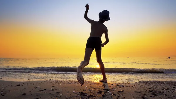 海の水の表面にスキップ石を投げ帽子を持つ少年のシルエット 鮮やかなオレンジ色の空と夏の休暇のコンセプト — ストック写真