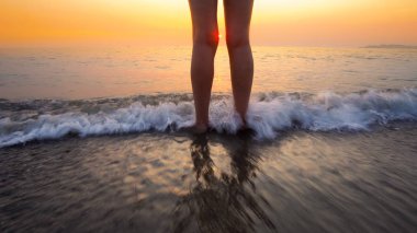 Sahilde gün batımında kadın ayaklarına su sıçrattı. Huzurlu ve rahatlatıcı okyanus suyu sahnesi güzel sahilde ayaklara sıçrıyor.