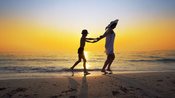 幸せな家族は海の海岸を歩き 日没時に波を蹴ります 母は白いドレスを着て帽子をかぶって — ストック写真