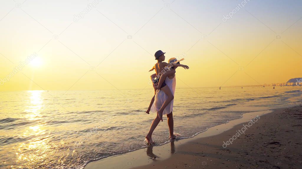 Family having fun on beach on travel vacation summer holidays. Young happy boy and mother doing playful joyful piggybacking ride outdoors at vibrant sunset