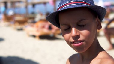 Young boy dazzled by summer sun on beach put on protection hat and smile to camera clipart