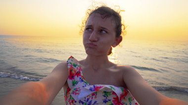 Bikinili güzel bir genç, arka planda yaz mevsiminin günbatımı ile komik selfie suratları yapar.