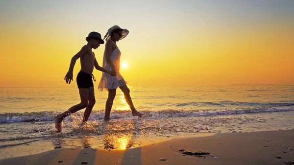 快乐的一家人在海滨散步 夕阳西下的浪花荡漾着浪花 妈妈穿着白色的衣服 两个人都有帽子 — 图库照片