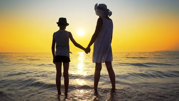 白いドレスと帽子と母と夕日を見てビーチに裸足で立っている帽子の息子は 海の波によってはねた 旅行の概念 — ストック写真
