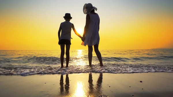 Family walk on beach to sea sunset and splash waves. Woman wear white dress and straw hat, boy has hat