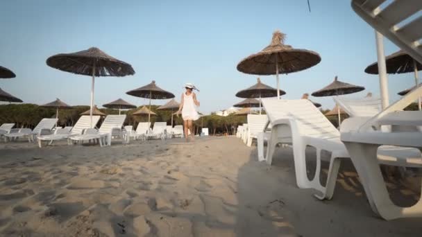白いドレスと帽子の幸せな笑顔の女性は わら傘とベッドで空の美しいビーチを歩いて風に吹かれた — ストック動画
