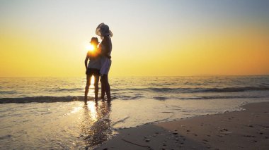 Aile günbatımı ve splash dalgalar deniz sahilde yürümek. Kadın beyaz elbise ve hasır şapka, şapka çocuk vardır