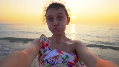 Bikinili güzel bir genç, arka planda yaz mevsiminin günbatımı ile komik selfie suratları yapar.