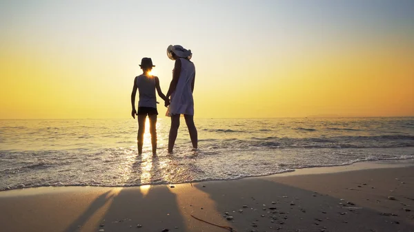 白いドレスと帽子と母と夕日を見てビーチに裸足で立っている帽子の息子は 海の波によってはねた 旅行の概念 — ストック写真