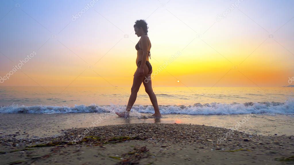 Fit woman walk on beach at sunset splashing water on sea waves