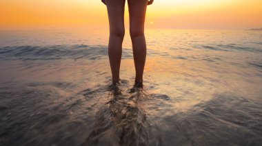 Sahilde gün batımında kadın ayaklarına su sıçrattı. Huzurlu ve rahatlatıcı okyanus suyu sahnesi güzel sahilde ayaklara sıçrıyor.