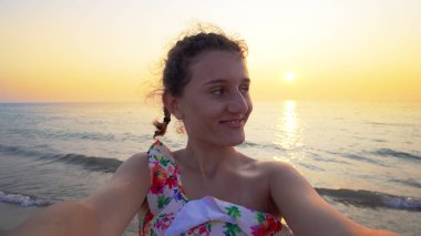 Bikinili güzel bir genç, arka planda yaz mevsiminin günbatımı ile komik selfie suratları yapar.