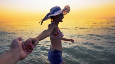 Günbatımında balayında romantik bir çift. Sahilde erkek arkadaşının elini tutarak yürüyen şapkalı, gülümseyen bir kız arkadaş. Yaz tatilinin tadını çıkaran bir çift.