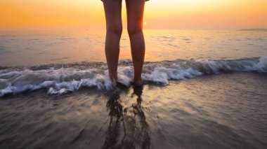 Sahilde gün batımında kadın ayaklarına su sıçrattı. Huzurlu ve rahatlatıcı okyanus suyu sahnesi güzel sahilde ayaklara sıçrıyor.