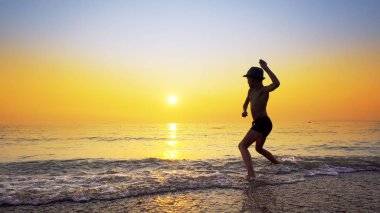 Şapkalı, taş atan, deniz suyu yüzeyinde zıplayan çocuk silueti. Canlı turuncu gökyüzü ile yaz tatili konsepti