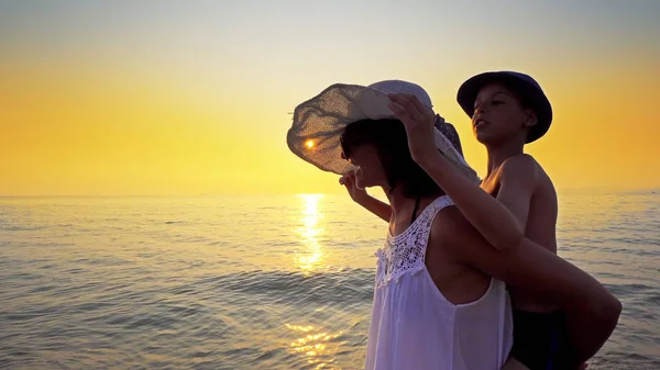 Mother gives her son a piggy back ride walking on empty ocean beach against sunset