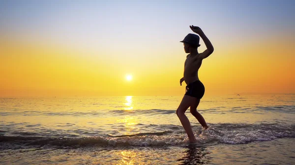 Silhouette of boy with hat throwing stones skipping on sea water surface. Summer vacation concept with vibrant orange sky